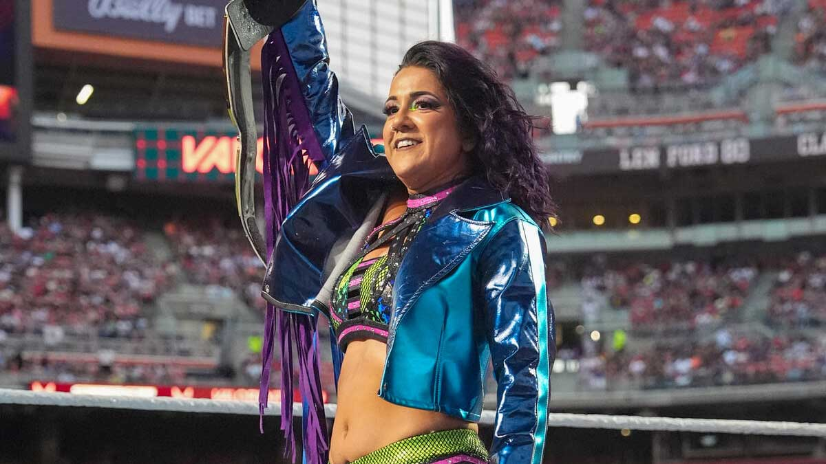 Bayley levanta el Campeonato Femenino de WWE mientras celebra sobre el ring ante miles de aficionados en un estadio durante un evento de la empresa.