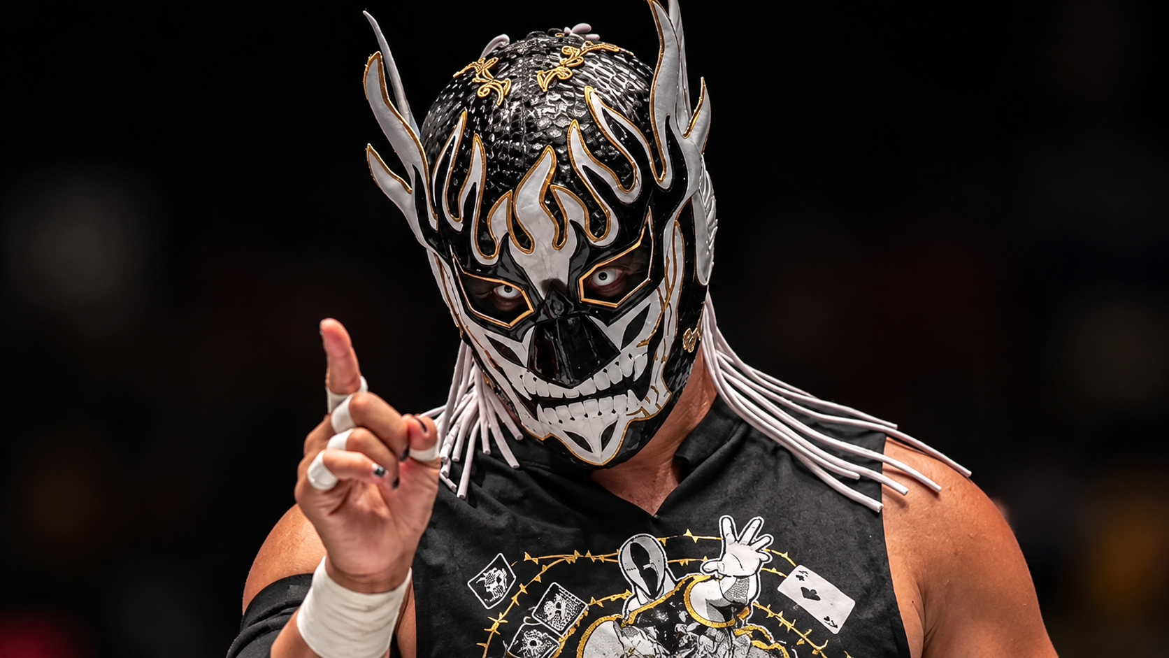 l Desperado, luchador de NJPW, aparece con su característica máscara negra, blanca y dorada, mientras levanta un dedo durante una presentación.