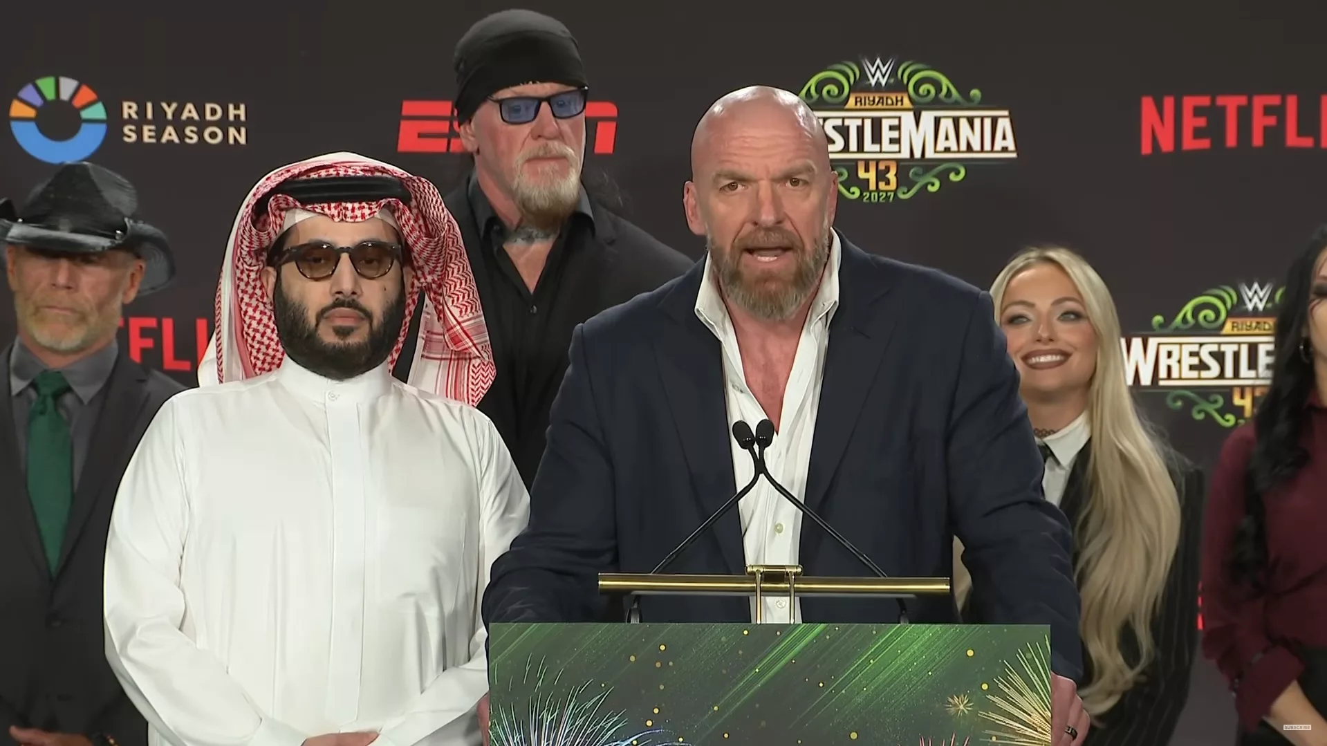 Triple H habla desde un podio junto a Turki Alalshikh, con The Undertaker y Liv Morgan detrás.