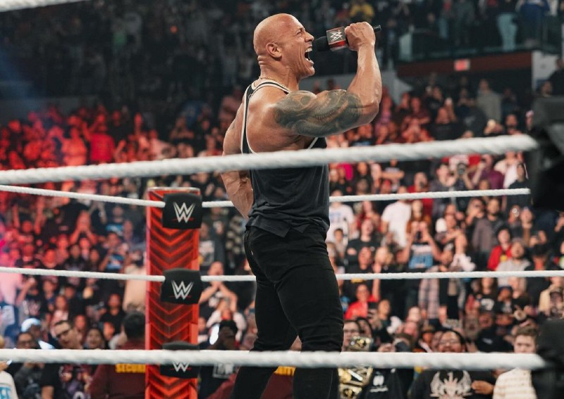 The Rock sostiene un micrófono de WWE dentro del ring durante una presentación en WWE Raw, frente al público.