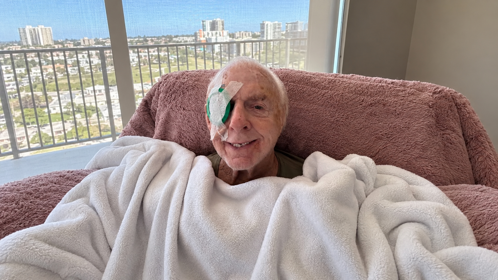 Ric Flair sonríe sentado, cubierto con una manta blanca y con un protector sobre un ojo tras sufrir una caída.