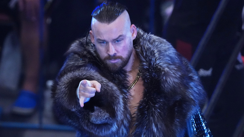 David Finlay, luchador de AEW y líder de The Dogs, señala hacia el frente mientras luce un abrigo negro de pelaje.